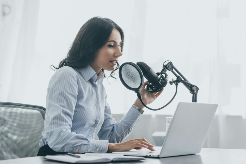 pretty radio host using laptop while speaking in microphone at workplace in broadcasting studio