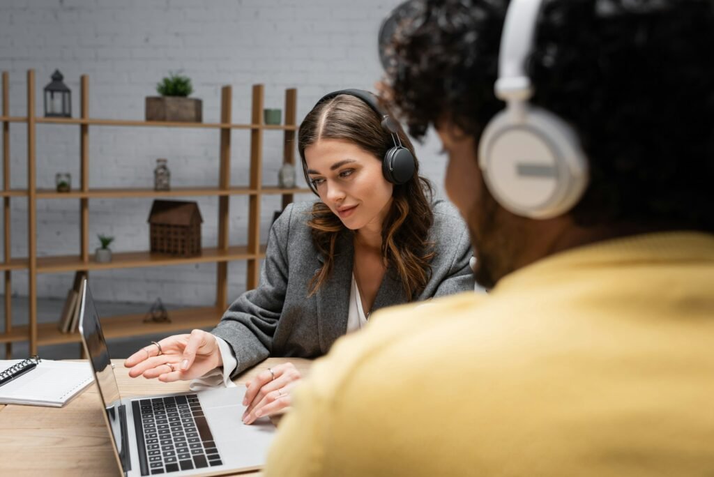 smiling brunette radio host in headphones and grey blazer pointing at laptop near notebook